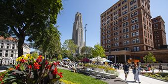 Cathedral of Learning and William Pitt Union surrounded by summer flowers with people walking outdoors.